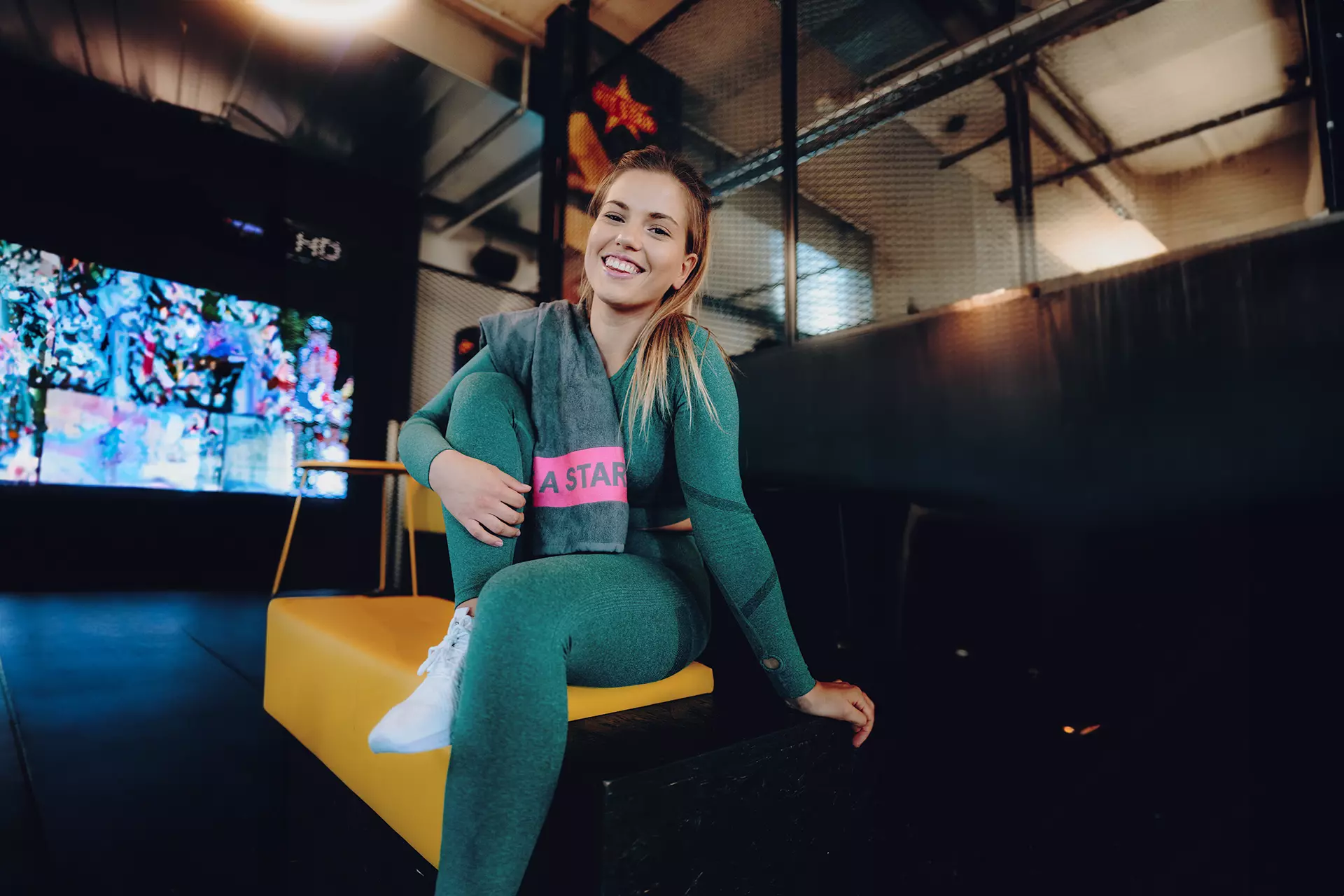 Frau mit blonden Haaren trägt ein grünes Sportoutfit mit angewinkelten Bein auf einer Sitzbank